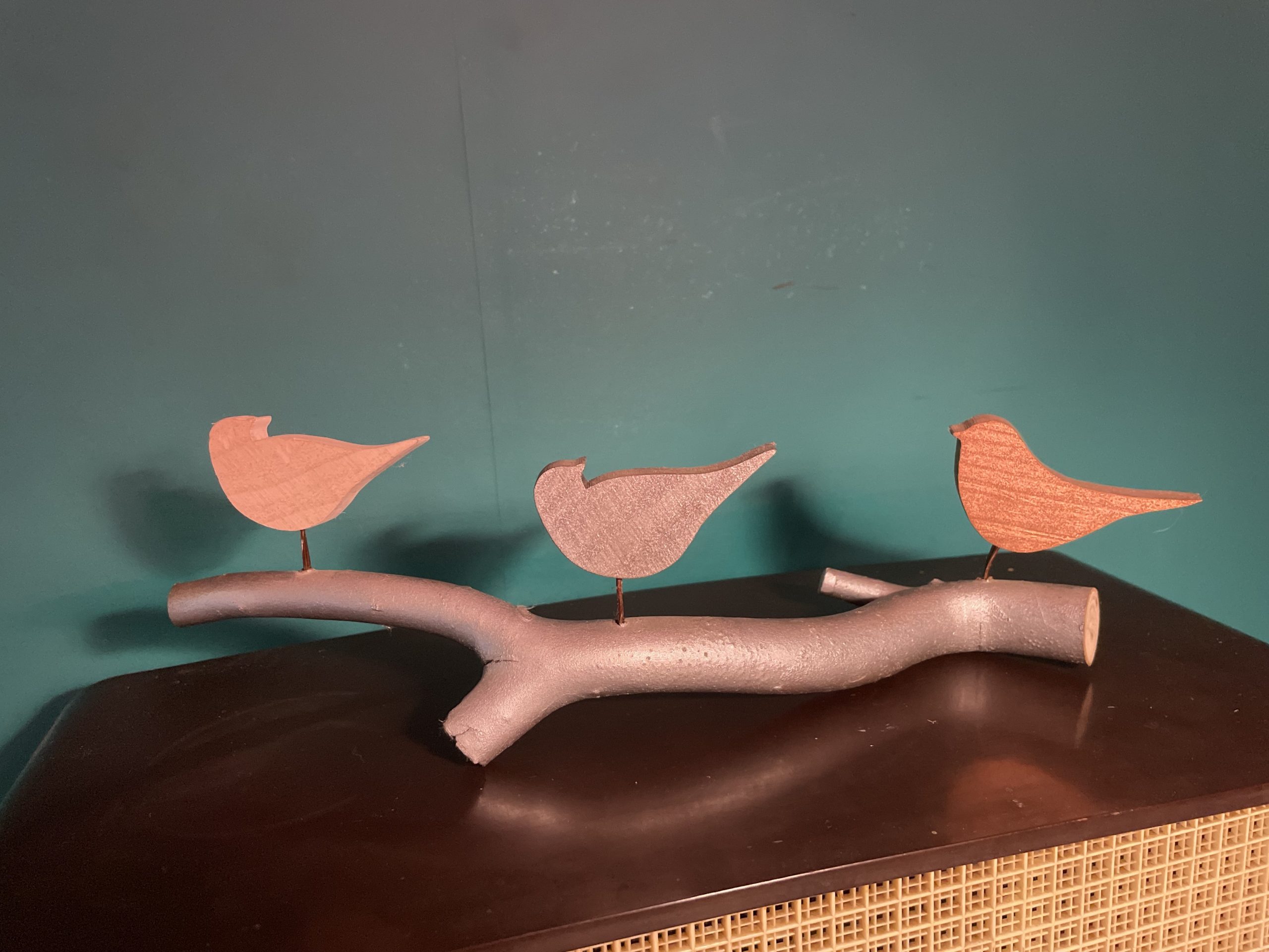 Handcrafted bird sculpture with three painted wooden birds on a silver-painted branch, made from reclaimed wood with copper wire legs. From the back.