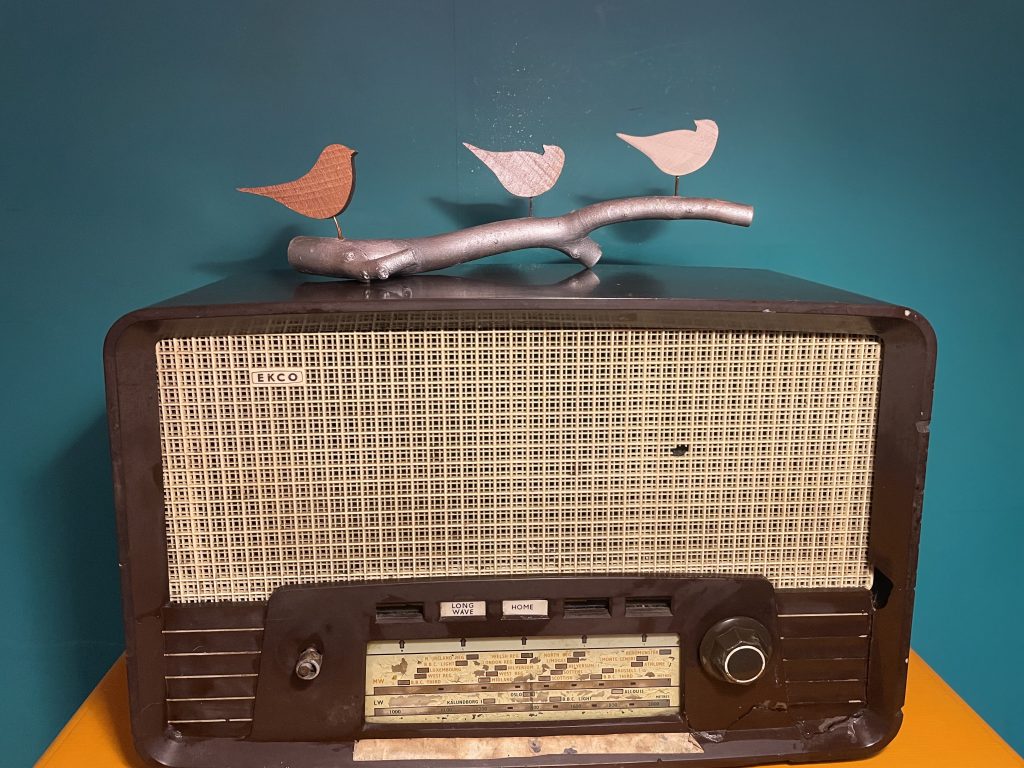 Handcrafted bird sculpture with three painted wooden birds on a silver-painted branch, made from reclaimed wood with copper wire legs. Sculpture on vintage wireless.